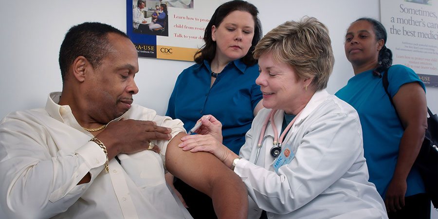 a doctor performing an injection on the left arm of a black man while two other women watching